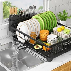 Puede incluir: Un escurreplatos negro con varios platos, cuencos y utensilios. El estante contiene platos blancos y verdes, vasos y cubiertos. Un fregadero y un grifo de acero inoxidable están en primer plano, con una ventana y vegetación en el fondo.