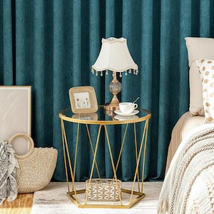 May include: A bedroom scene featuring a gold side table with a glass top, a lamp, and a framed photo. A woven tote bag and a framed abstract art piece are on the left. A bed with a patterned pillow and a blanket is on the right. Teal curtains form the backdrop.