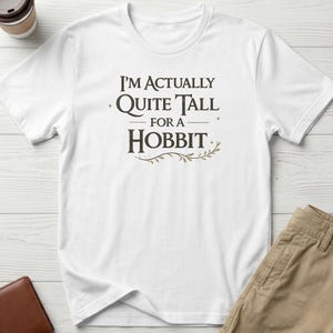 May include: A white short-sleeved t-shirt featuring the dark brown text "I'm Actually Quite Tall For A Hobbit" centred on the chest. The design includes small star accents and a decorative leafy vine graphic underneath the word Hobbit.