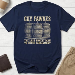 May include: A navy blue short-sleeved t-shirt featuring a graphic print of three wooden barrels with fuses. Above the barrels is the text GUY FAWKES, and below the barrels is the text THE LAST HONEST MAN TO ENTER PARLIAMENT in a light beige colour.