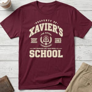 May include: A maroon short-sleeved t-shirt featuring a cream-coloured graphic print that reads Property of Xavier's School for Gifted Youngsters, Est. 1963, 1407 Graymalkin Lane, Salem Center, N.Y., with an open book and DNA strand icon.