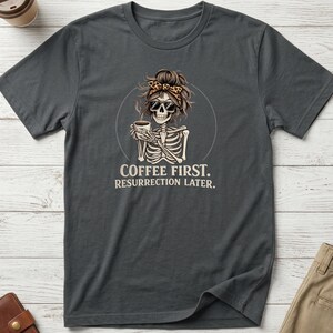 May include: A charcoal grey short-sleeve t-shirt featuring a graphic of a skeleton wearing sunglasses and a leopard print headband, holding a coffee mug. The text below the skeleton reads, COFFEE FIRST. RESURRECTION LATER. The shirt has a crew neckline.
