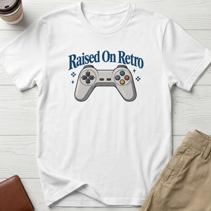 May include: A white short-sleeved t-shirt featuring a graphic of a grey video game controller centred on the chest. Above the controller, the text "Raised On Retro" is printed in a dark blue font, accompanied by small blue star and pixel icons.