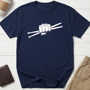 May include: A navy blue short-sleeved t-shirt featuring a white graphic print centred on the chest. The graphic depicts a clenched fist holding two drumsticks in a horizontal position, rendered in a clean, high-contrast line art style.