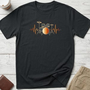 May include: A black short-sleeved t-shirt featuring a graphic print of a drum kit centred on the chest. The drum kit is integrated into a horizontal heartbeat line design, coloured in shades of orange, yellow, and brown with a retro aesthetic.
