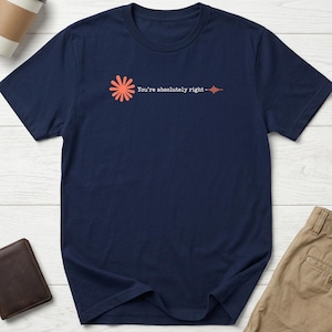 May include: A navy blue short-sleeved t-shirt featuring a coral-coloured sunburst icon on the left chest. Next to the icon, the text "You're absolutely right" is printed in white, followed by a small horizontal sound wave graphic in coral.
