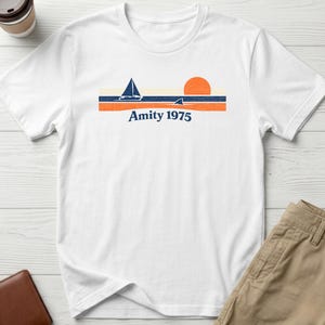 May include: White t-shirt with a nautical design. It features a sailboat, sun, and shark fin above the text "Amity 1975". The design includes blue and orange horizontal stripes. The shirt is made of a soft material.