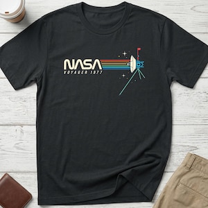 May include: A black short-sleeved t-shirt featuring a graphic print on the chest. The design includes the white text NASA VOYAGER 1977, a horizontal multi-coloured striped line, and a white illustration of the Voyager space probe with stars.