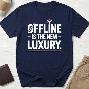 May include: A navy blue t-shirt with the white text "OFFLINE - IS THE NEW - LUXURY." The letter "i" in "offline" is replaced with a wifi symbol. The shirt is on a light wood surface, with a brown wallet and beige trousers visible.
