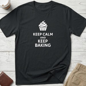 May include: Black t-shirt featuring a white cupcake design and the words "KEEP CALM and KEEP BAKING". A comfortable tee for those who enjoy baking, made from a soft fabric.