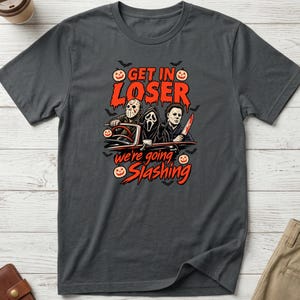 May include: A charcoal grey short-sleeve t-shirt featuring a graphic of three horror movie characters in a red convertible. The text reads GET IN LOSER we're going SLASHING, surrounded by small orange jack-o'-lanterns and black bat silhouettes.