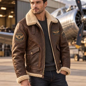 May include: A brown leather bomber jacket with a cream-coloured faux shearling collar and cuffs. The jacket features a front zip, two flap pockets, and circular patches on the sleeves and chest with text including Higher Further Together and Skybound Aviation.