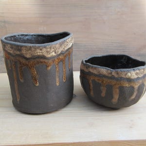 May include: Two handmade ceramic vessels with a dark charcoal matte finish. Both pieces feature a textured, uneven rim and a decorative golden-brown glaze that drips down the sides, creating a distinct contrast against the dark clay body.