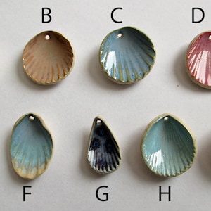 May include: Nine ceramic shell-shaped pendants labelled A through I, each featuring a small hole for hanging. The collection includes various glazed finishes in shades of blue, tan, pink, and dark purple, with visible ridges and scalloped edges.