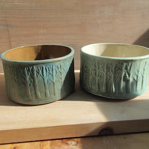 May include: Two ceramic bowls with a forest tree design. The bowls are oval-shaped with a teal exterior and a tan interior. The tree design is carved into the exterior of the bowls. The bowls are sitting on a wooden surface.