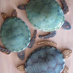 May include: Three decorative turtle-shaped wall plaques featuring smooth, glossy, green-toned shells with a mottled finish. Each turtle has textured, brown-coloured flippers and a head with a distinct, segmented, stone-like pattern on the surface.