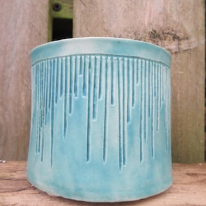 May include: A turquoise ceramic plant pot with a cylindrical shape and textured vertical lines. The lines give a dripping effect around the top. The pot sits on a wooden surface, with a wooden background.