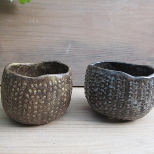 May include: Two handmade ceramic bowls with textured surfaces. One bowl is brown with yellow accents, and the other is dark grey. The bowls have an organic, irregular shape, suitable for serving or display.