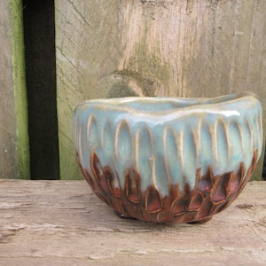 May include: A handcrafted ceramic bowl with a distinctive design. The bowl has a light blue glaze at the top, fading into a textured, brown base. It rests on a weathered wooden surface.