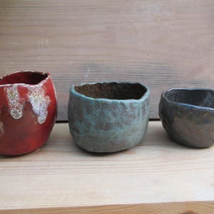 May include: Three small, hand-formed ceramic bowls with irregular, organic shapes. The left bowl features a glossy red glaze with white textured accents. The centre bowl has a matte, mottled teal finish. The right bowl is a dark, metallic charcoal grey.