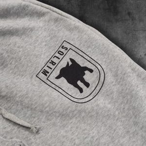 May include: Close-up of gray sweatpants with a drawstring. A black and white logo is printed on the upper leg. The logo is a shield shape with the word "SOLRIM" and a silhouette of an animal.
