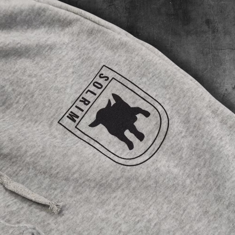 May include: Close-up of gray sweatpants with a drawstring. A black and white logo is printed on the upper leg. The logo is a shield shape with the word "SOLRIM" and a silhouette of an animal.