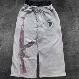 May include: Light gray sweatpants with a drawstring waist and a large graphic of an AK-47 rifle printed on the left leg. The right leg features a black emblem with the text "WIRLOS". A pair of black sweatpants are folded on top.