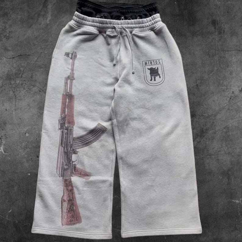 May include: Light gray sweatpants with a drawstring waist and a large graphic of an AK-47 rifle printed on the left leg. The right leg features a black emblem with the text "WIRLOS". A pair of black sweatpants are folded on top.