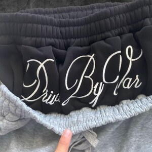 May include: Close-up of gray sweatpants with a drawstring and black sweatpants peeking out from the top. The black sweatpants have the words "Drive By Car" embroidered in white cursive lettering.