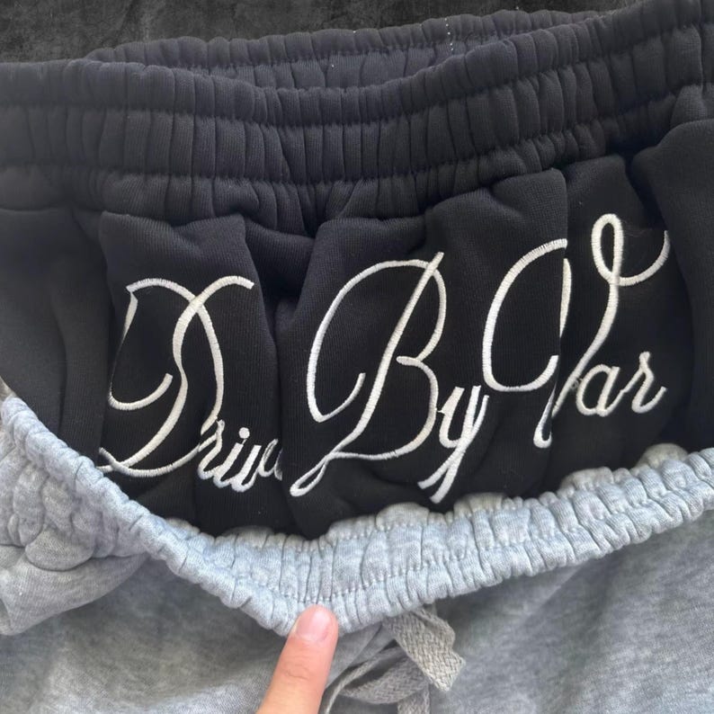 May include: Close-up of gray sweatpants with a drawstring and black sweatpants peeking out from the top. The black sweatpants have the words "Drive By Car" embroidered in white cursive lettering.