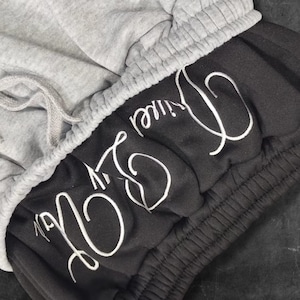 May include: Close-up of gray and black sweat shorts. The shorts have a gray drawstring waistband and the words "Live Divine" embroidered in white script on the black fabric. The shorts are made of a soft material.