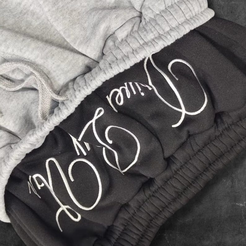 May include: Close-up of gray and black sweat shorts. The shorts have a gray drawstring waistband and the words "Live Divine" embroidered in white script on the black fabric. The shorts are made of a soft material.