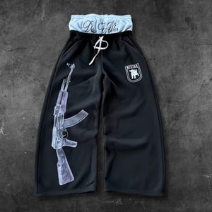 May include: Black sweatpants with a white drawstring and a light blue waistband. The left leg features a graphic of a transparent AK-47 rifle. A white emblem with a bull is on the right leg. The waistband has the text "D.W.S."