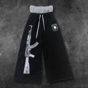 May include: Black wide-leg sweatpants with a gray waistband and white drawstring. A detailed graphic of a rifle is printed on the left leg. A white emblem with a bull is on the right leg. The word "Rebel" is printed on the waistband.