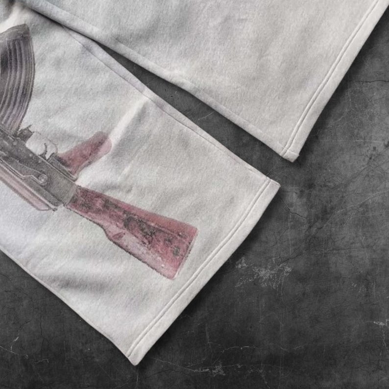 May include: Light gray sweatpants featuring a detailed graphic of an AK-47 assault rifle in shades of red and gray. The pants have a relaxed fit with a stitched hem. The image is a close-up, showing the fabric's texture.