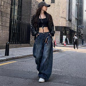 Pode incluir: Uma pessoa vestindo um top preto, uma jaqueta de couro preta e jeans de perna larga de lavagem escura. A pessoa est&aacute; usando um bon&eacute; de beisebol e t&ecirc;nis brancos. A roupa &eacute; usada em uma rua da cidade.
