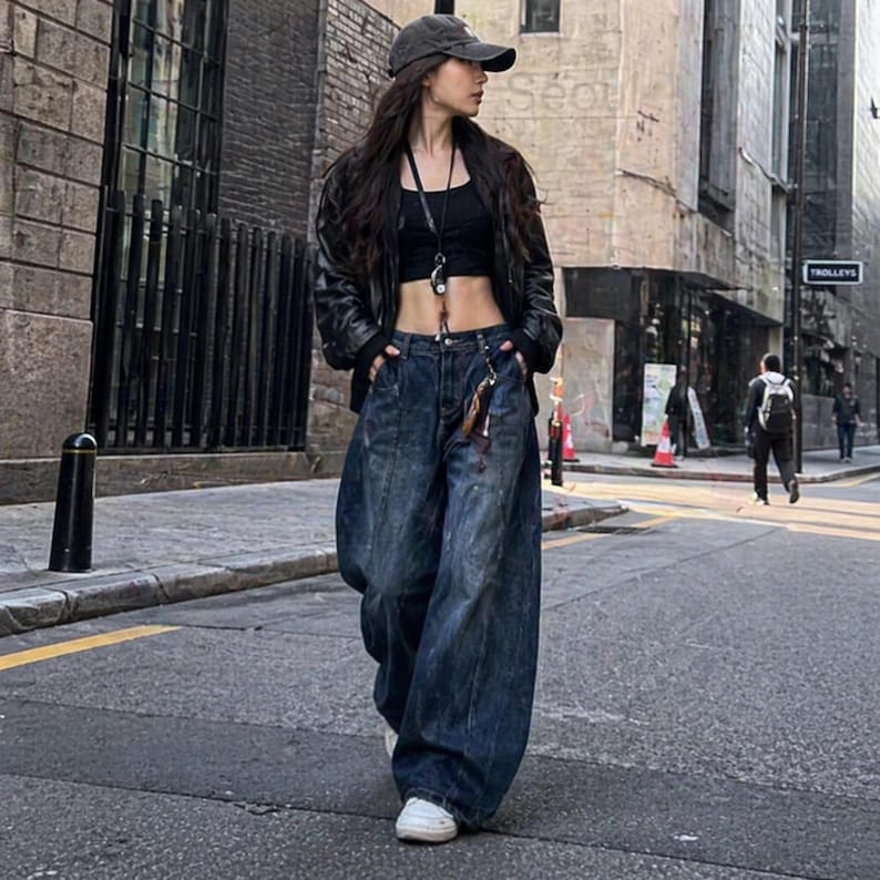 Pode incluir: Uma pessoa vestindo um top preto, uma jaqueta de couro preta e jeans de perna larga de lavagem escura. A pessoa est&aacute; usando um bon&eacute; de beisebol e t&ecirc;nis brancos. A roupa &eacute; usada em uma rua da cidade.