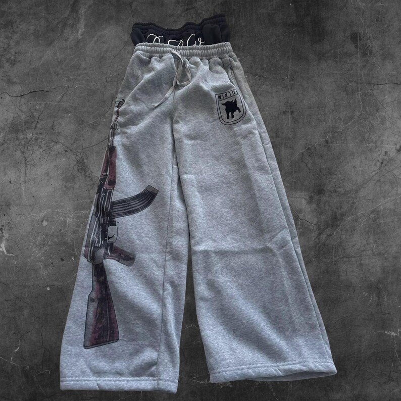 May include: Gray sweatpants with a drawstring waist and a large graphic of an AK-47 rifle on the left leg. The right leg features a small patch with a bull graphic and the text "H1R10". A black top is visible at the top.