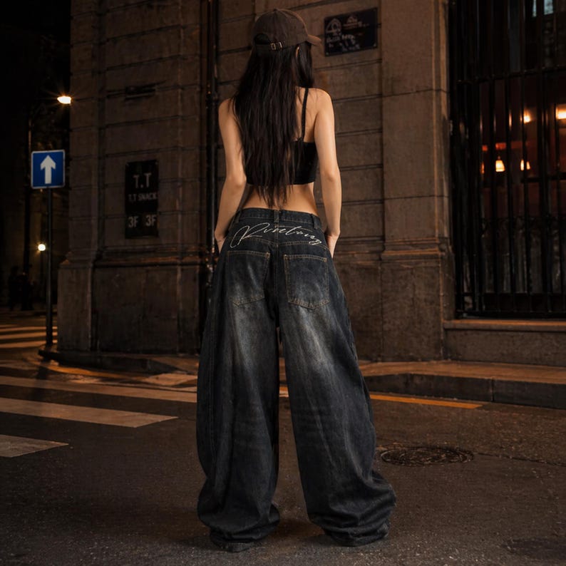 Pode incluir: Jeans de ganga de lavagem escura com pernas largas e a palavra "Pinebong" bordada nos bolsos traseiros. Os jeans t&ecirc;m um corte descontra&iacute;do e s&atilde;o usados com um top curto preto e um bon&eacute; de beisebol castanho.