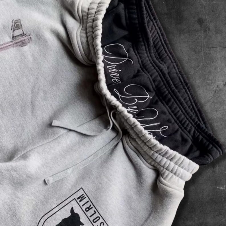 May include: Close-up of gray sweatpants with a black waistband. The gray portion has an embroidered design and a drawstring. The black waistband features the cursive text "Drive By Mike". A logo is visible on the gray portion.