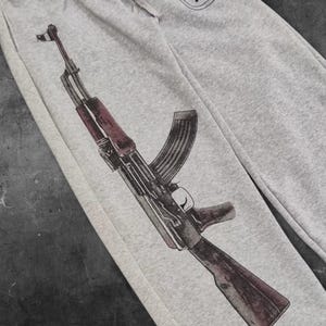 May include: Light gray sweatpants featuring a detailed illustration of an AK-47 assault rifle in shades of brown, black, and red. The graphic is printed on the left leg of the pants. The pants are made of a soft, comfortable material.