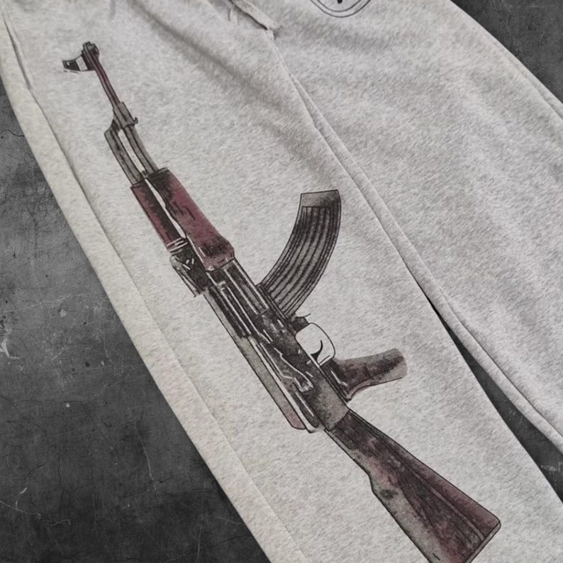 May include: Light gray sweatpants featuring a detailed illustration of an AK-47 assault rifle in shades of brown, black, and red. The graphic is printed on the left leg of the pants. The pants are made of a soft, comfortable material.