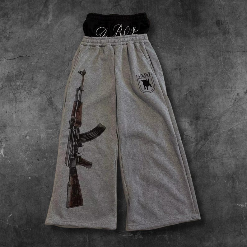 May include: Gray sweatpants with a large graphic of an AK-47 rifle on the left leg. The right leg features a black emblem with a bull logo and the word "NINTOS". A black top with the text "A.B.C" is visible at the top.