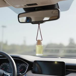 Puede incluir: Un ambientador para coche con tapa de madera y botella de vidrio transparente llena de líquido amarillo, colgado del espejo retrovisor. Se ve el interior del coche.