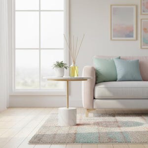 Pode incluir: Uma pequena mesa lateral redonda com um tampo de mármore branco e uma haste e base de metal dourado. Uma pequena planta em vaso e uma garrafa difusora de vidro com juncos estão sobre a mesa. Um sofá rosa claro com duas almofadas está ao fundo.