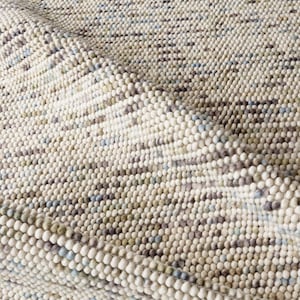 May include: Close-up of a textured, off-white rug with a pattern of small, raised, spherical elements. The rug has a mix of neutral colours, including beige, grey, and hints of blue and green. The rug's texture is visible, with a folded section.