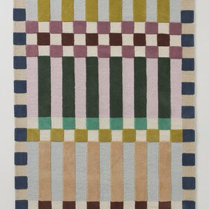 May include: Rectangular rug with a geometric pattern. The rug features vertical stripes in shades of green, pink, tan, and blue, interspersed with checkered sections in brown, white, and purple. The border is a series of blue squares.