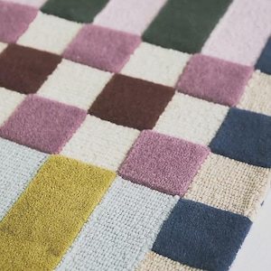 May include: A patterned rug with a mix of colours. The design includes green and pink stripes, and a checkerboard section with brown, purple, white, yellow, blue, and beige squares. The rug is made of a soft material.