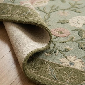 May include: A sage green area rug featuring a floral pattern with light pink and white blossoms. The rug has a darker olive green border with a leaf motif. One corner is folded back, revealing the light-colored, woven fabric backing.