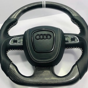 Könnte beinhalten: Schwarzes Lenkrad aus Kohlefaser mit weißen Nähten und einem Audi-Logo. Das Lenkrad verfügt über einen perforierten Ledergriff und Bedientasten. Ein weißer Streifen akzentuiert die Oberseite des Lenkrads.
